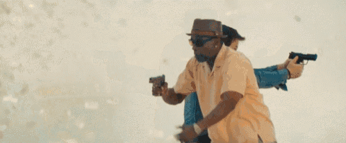 a man in a hat and sunglasses is holding two guns in front of a woman .