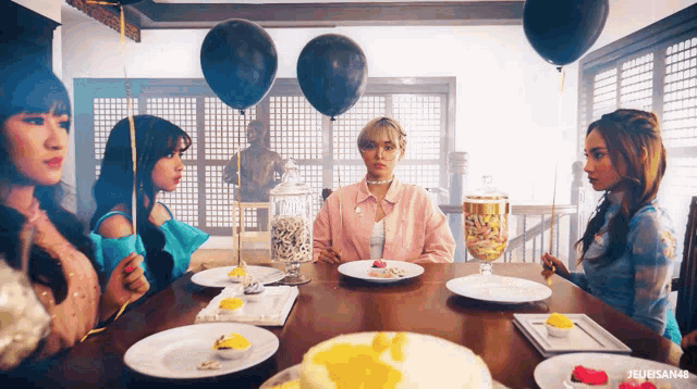 a group of girls are sitting around a table with plates of food and balloons hanging from the ceiling ..