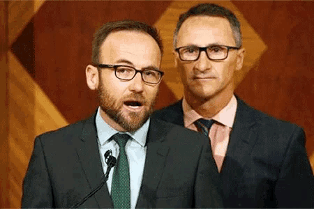 a man with glasses and a beard is speaking into a microphone while another man stands behind him