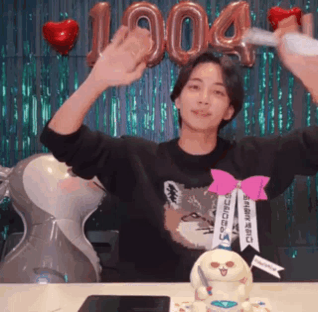 a man wearing a black shirt with a cat on it is sitting at a table with a cake and balloons that say 1904