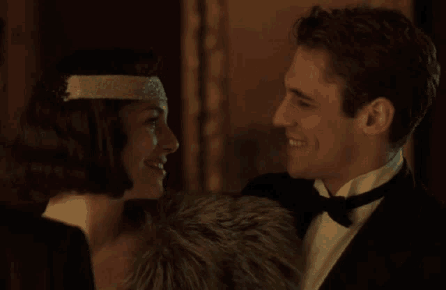 a man in a tuxedo and a woman in a headband are smiling at each other