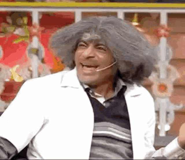 a man wearing a gray wig and a white coat is sitting in front of a microphone and smiling .