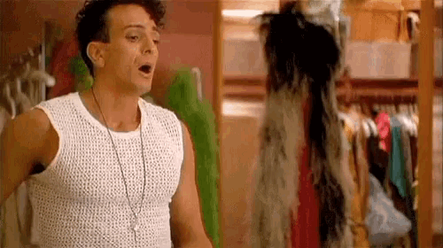 a man wearing a white tank top and a necklace is standing in a closet with his mouth open .
