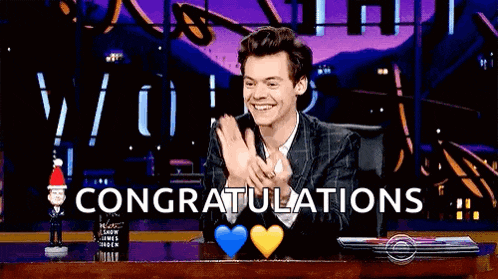 harry styles is smiling and clapping while sitting at a table with congratulations written on the screen .