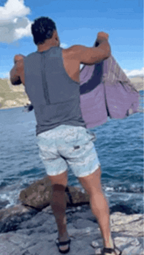 a man in a tank top and shorts is standing on a rock near the ocean .