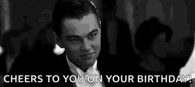 a man in a tuxedo is holding a glass in front of his face and says `` cheers to you on your birthday '' .