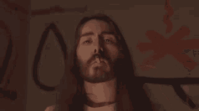 a man with long hair and a beard is standing in front of a wall with graffiti on it including the letters z and x