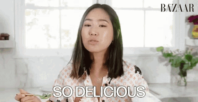 a woman is sitting in a kitchen eating food with chopsticks and says `` so delicious '' .