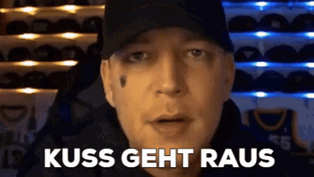 a man wearing a black hat is standing in front of a wall of hats and says kiss geht raus .