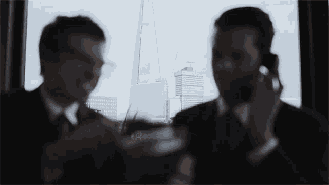 two men are talking on their cell phones in front of a window with a skyscraper in the background