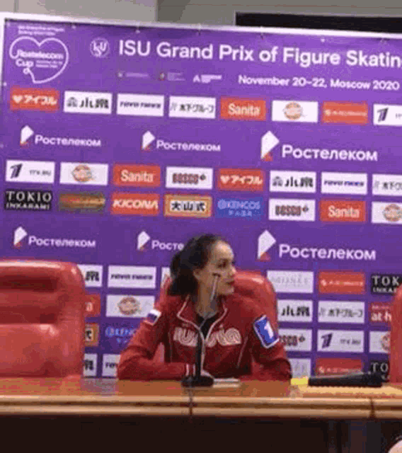 a woman is sitting at a table talking into a microphone in front of a purple sign that says isu grand prix