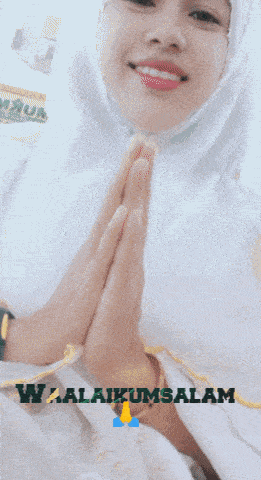 a woman wearing a white hijab with her hands folded in front of a sign that says ' selamat '