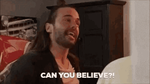 a man with long hair and a beard is talking to another man in a room .
