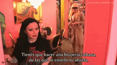 two women are standing in a pink room and one of them says " tienes que hacer una brujeria cubana
