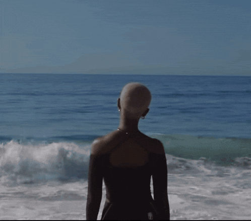 a woman with a shaved head is standing on the beach looking at the ocean