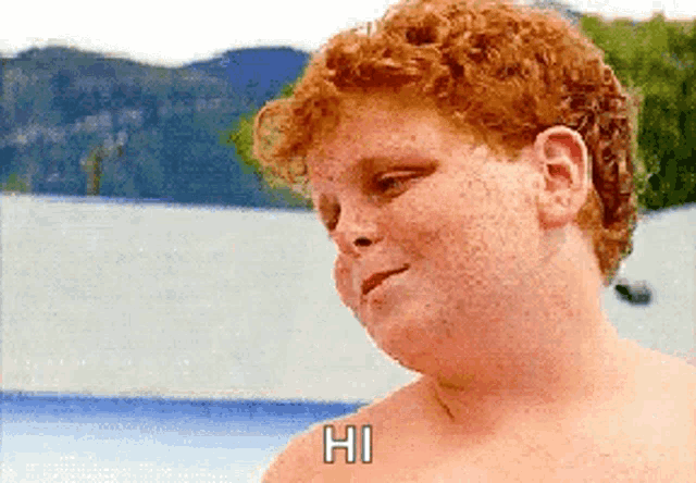a shirtless red haired boy says hi in front of a lake