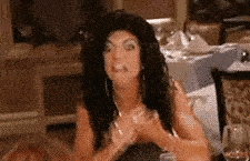 a woman is sitting at a table with a glass of wine and making a face .