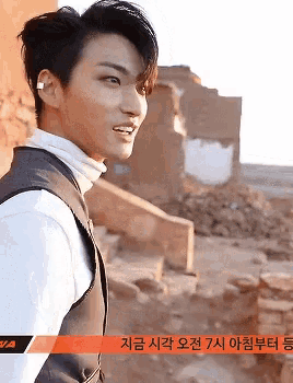 a man wearing a white turtleneck and a black vest stands in front of a brick wall with foreign writing on it