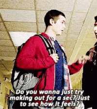 a man in a red jacket is talking to another man in a hallway while holding a backpack .