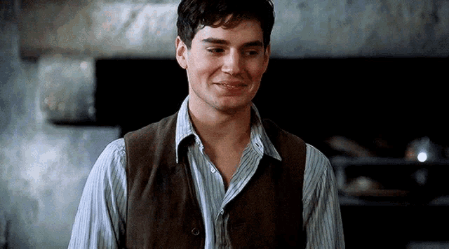 a man wearing a vest and a striped shirt is smiling while standing in front of a fireplace .