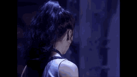 a woman with purple hair is standing in a dark room and looking at the camera .