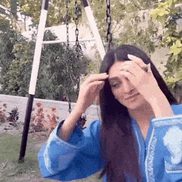 a woman in a blue shirt is sitting on a swing adjusting her hair