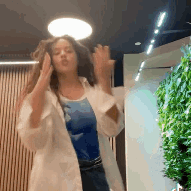a woman in a white coat is dancing in front of a wall of plants