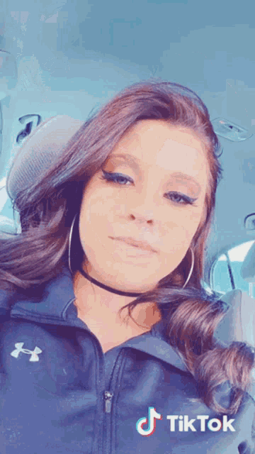a woman wearing a blue under armour jacket is sitting in a car with a tiktok sticker on her face