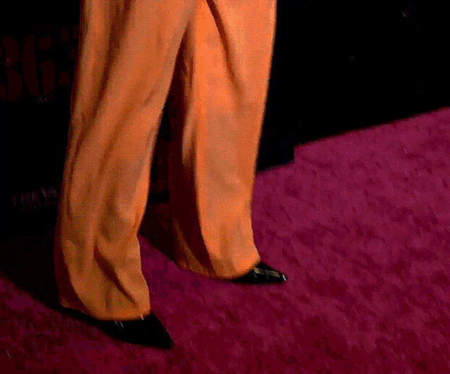 a person wearing orange pants and black shoes walking on a pink carpet