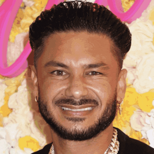 a man with a beard and earrings smiles in front of a pink sign