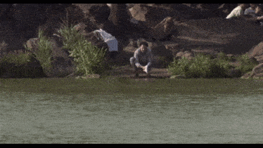 a man is squatting on the shore of a lake