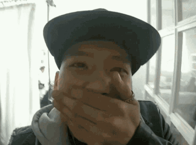 a man in a hat is covering his mouth with his hands .