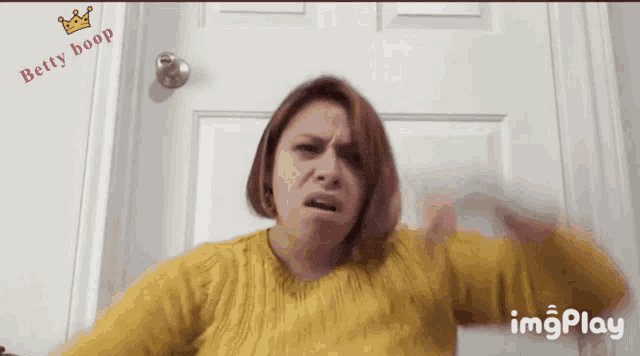 a woman in a yellow sweater is making a funny face in front of a door with the words betty boop on it