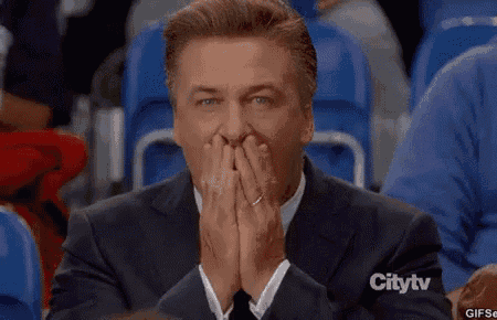 a man in a suit and tie is sitting in a stadium and covering his mouth with his hands and says " you 're "