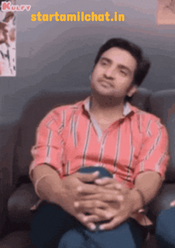 a man in a red and white striped shirt sits on a couch with his hands folded