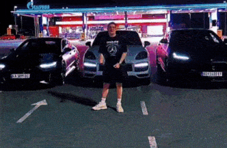 a man in a mercedes shirt is standing in front of three cars in a parking lot .