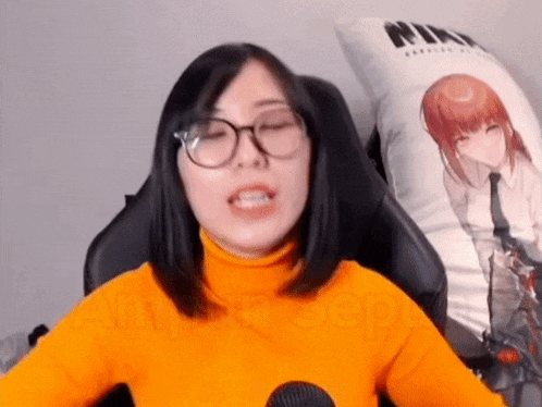 a woman wearing glasses and an orange turtleneck is sitting in a chair with a pillow in the background .