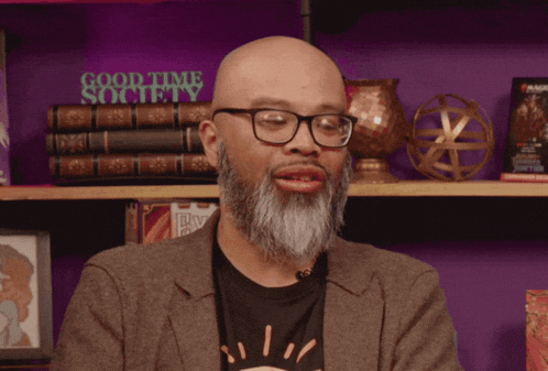 a bald man with glasses and a beard stands in front of a shelf with books and a sign that says good time society