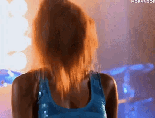 a woman in a blue tank top stands in front of a drum set with the word morangos on the bottom right