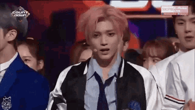 a man with pink hair and a tie is standing on a stage in front of a crowd of people .