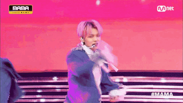 a man with pink hair is singing into a microphone on a stage with a pink background .