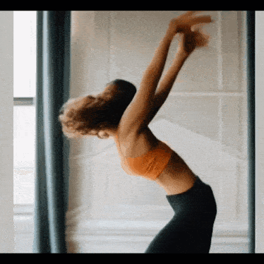 a woman in an orange top and black pants is doing yoga in front of a window