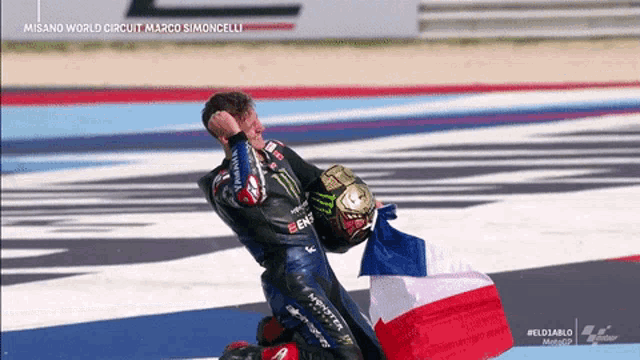 a motorcycle racer is kneeling down with a french flag in front of him with misano world circuit in the background