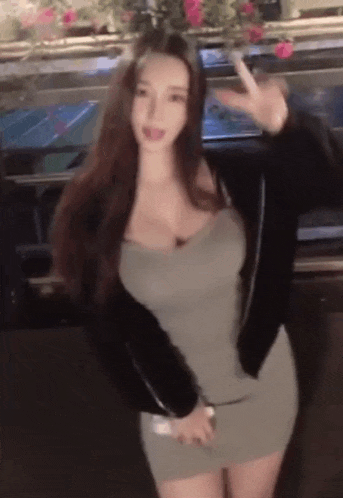 a woman in a grey dress and black jacket is standing in front of a car and waving .