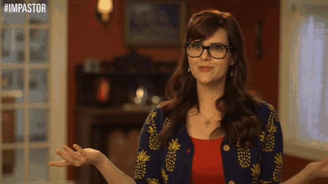 a woman wearing glasses and a blue cardigan with pineapples on it stands in front of a sign that says #impastor
