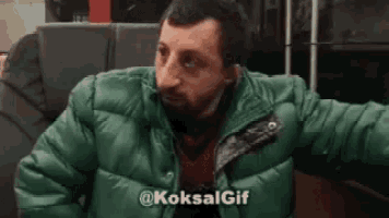 a man with a beard is wearing a green jacket and sitting on a train .