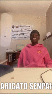 a man in a pink hoodie is sitting at a desk with the words " arigato senpai " written below him