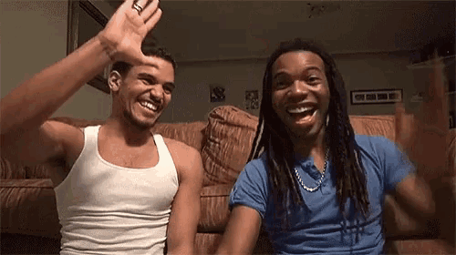 two men are sitting on a couch laughing and waving at the camera