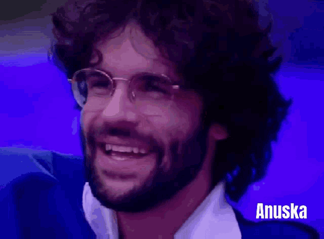 a man with glasses and a beard is smiling in front of a blue background that says anuska