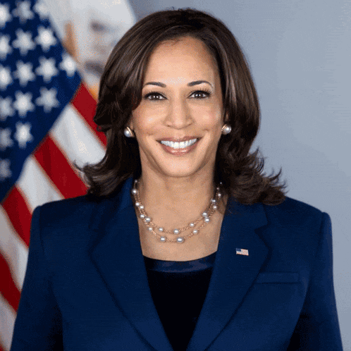 a woman in a blue suit and pearl necklace smiles in front of an american flag
